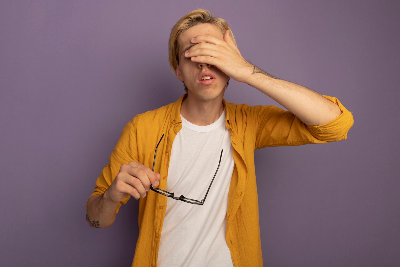 tired young blonde guy wearing yellow t-shirt holding glasses and covered eyes with hand isolated on purple background