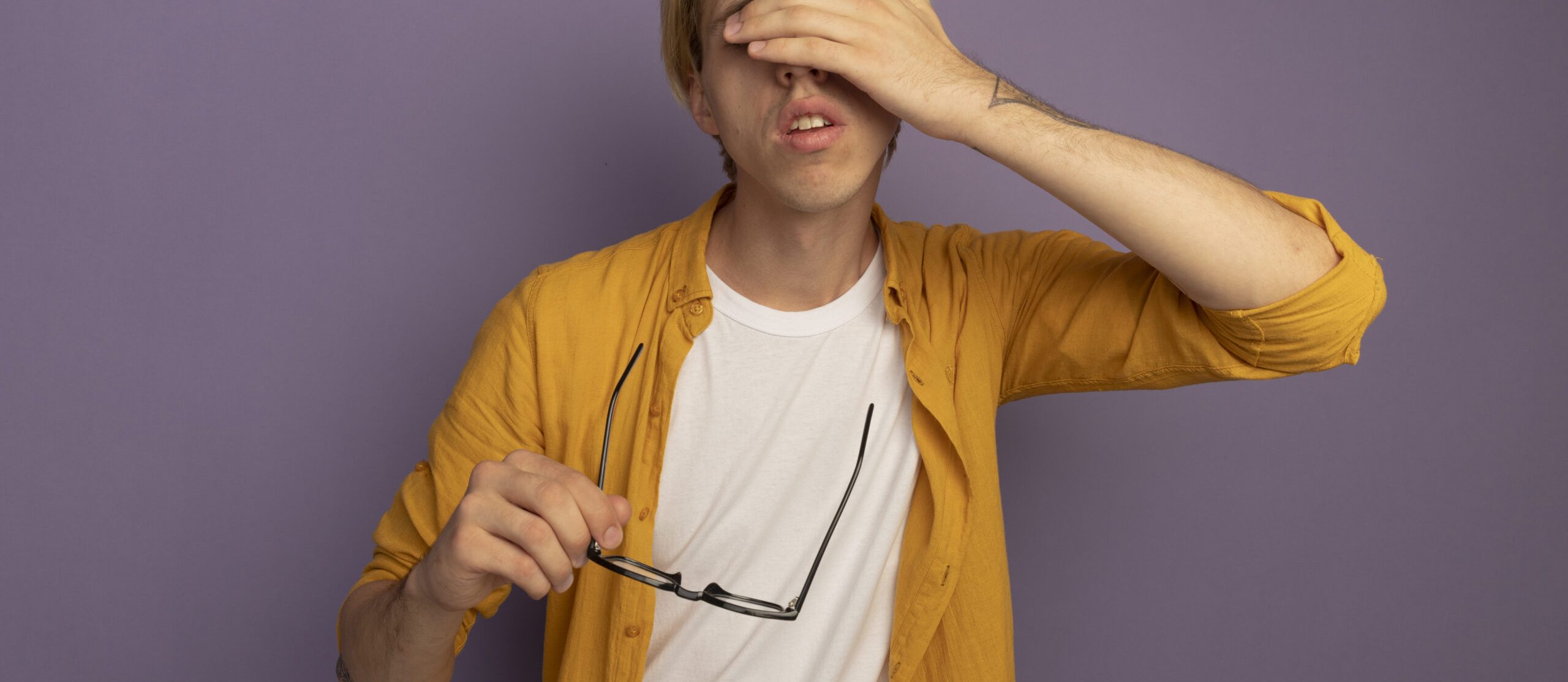 tired young blonde guy wearing yellow t-shirt holding glasses and covered eyes with hand isolated on purple background